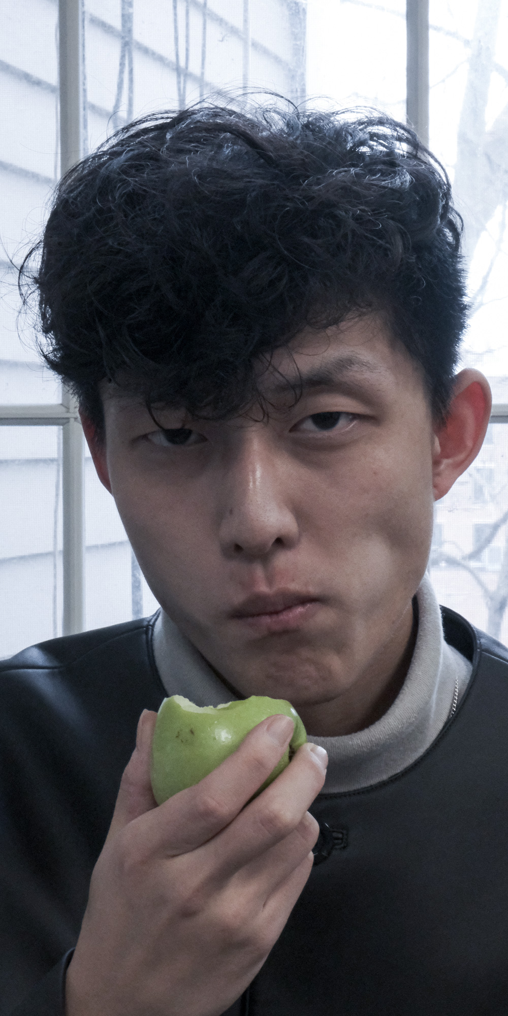 Young man with curly dark hair eating a green apple indoors near a window.