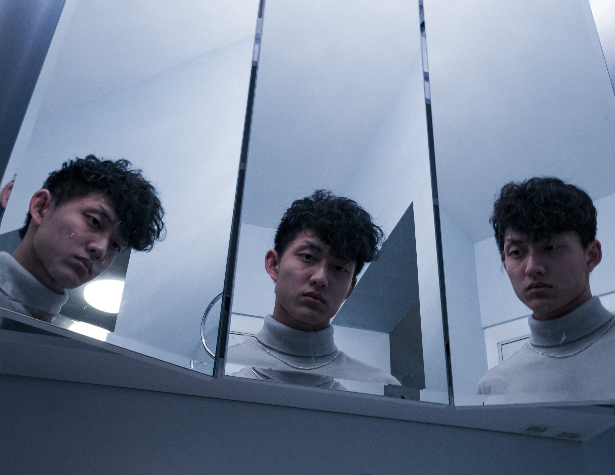 Young man with curly hair wearing a light turtleneck looking serious in three angled mirrors.
