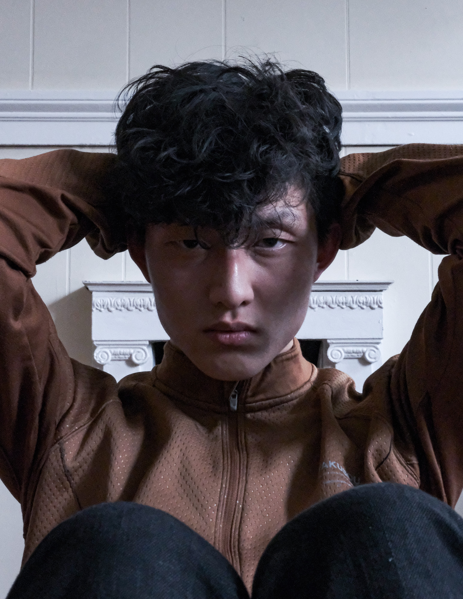 Young person with curly black hair wearing a brown jacket, sitting with hands behind head against a white wall with decorative molding.