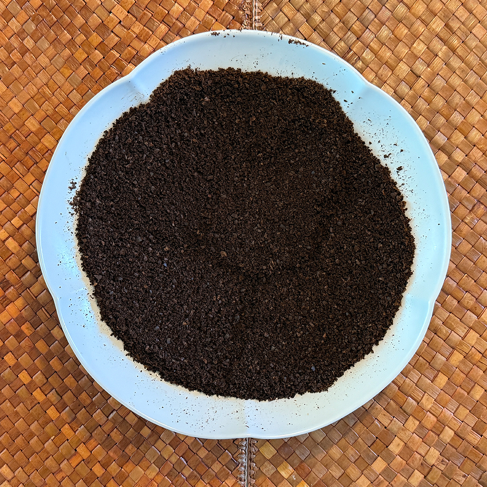 White scalloped plate filled with dark brown coffee grounds on a woven mat surface.