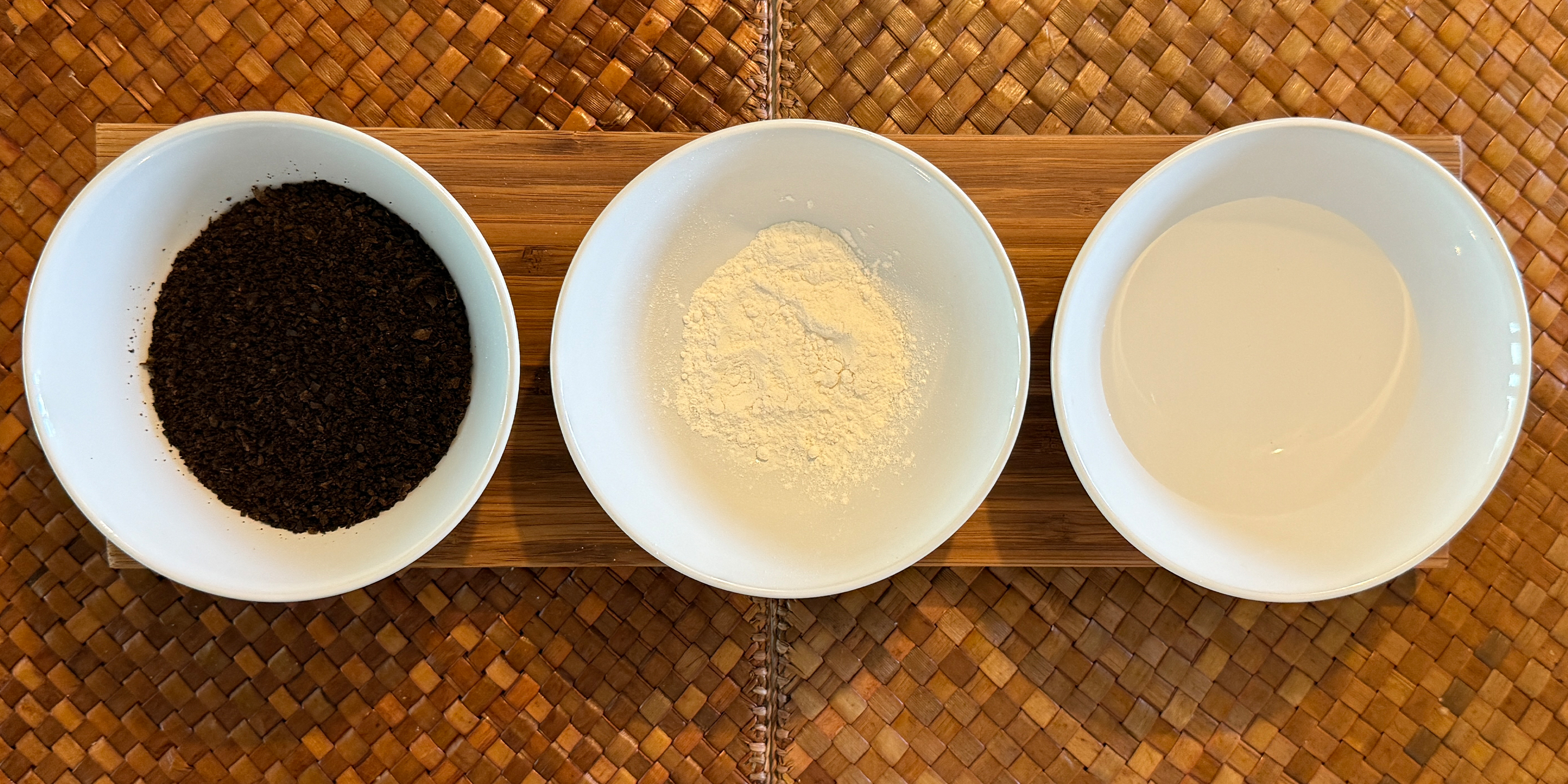 Top view of three white bowls on a wooden tray containing ground coffee, powdered creamer, and milk.