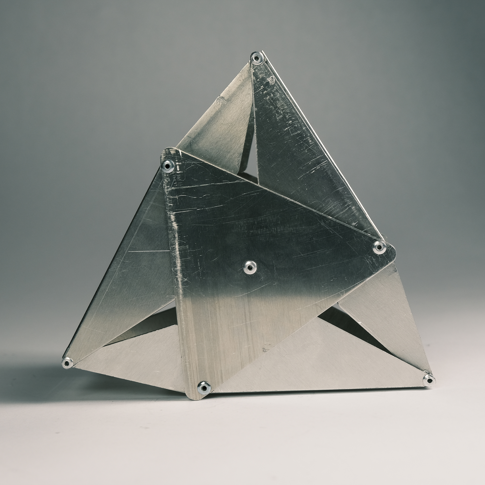 Metal geometric sculpture composed of triangular plates bolted together on a light surface with a gray background.
