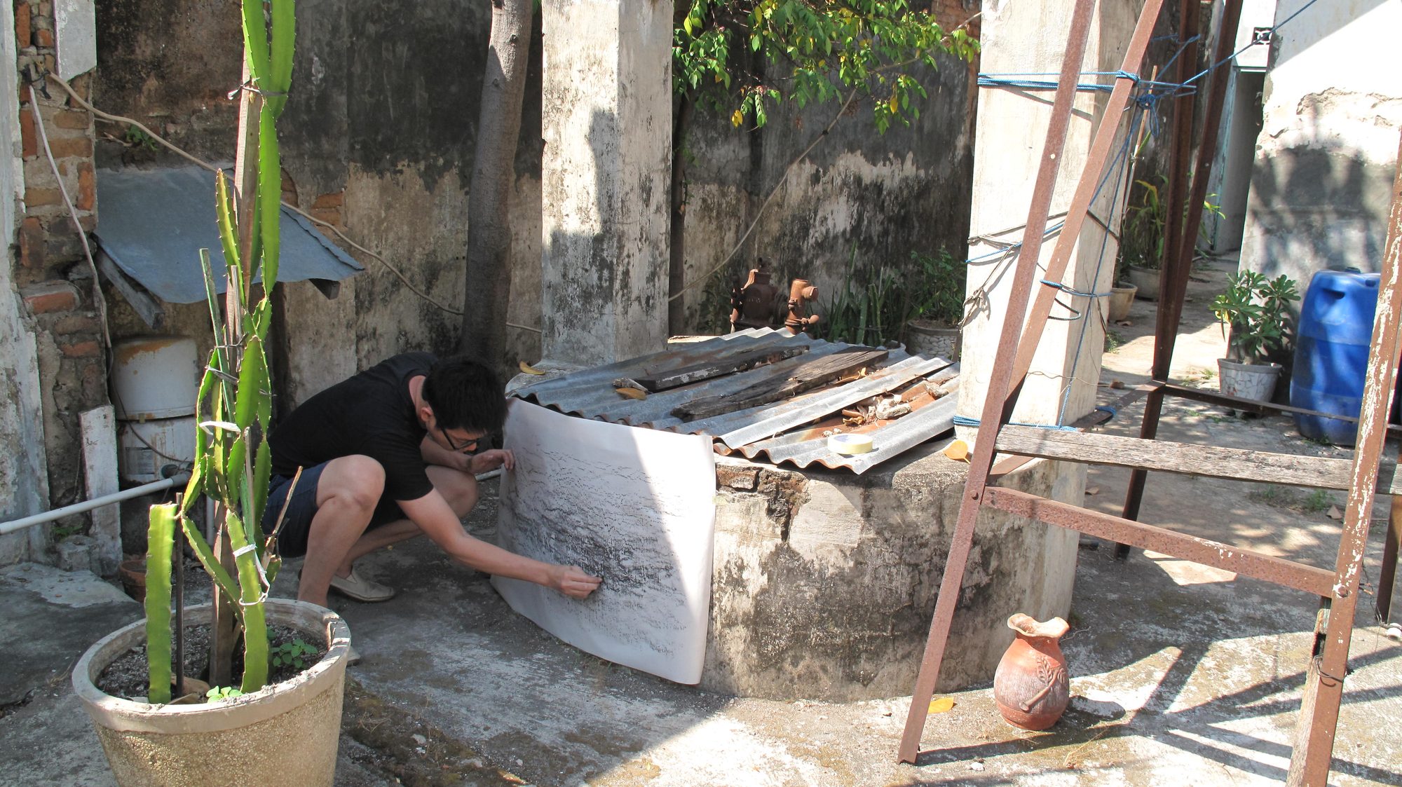 Boedi crouching to make a frottage in the courtyard of his childhood house