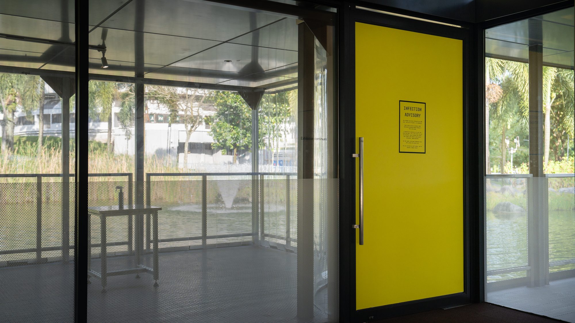 A glass room by a lake with a yellow door bearing an Infection Advisory notice