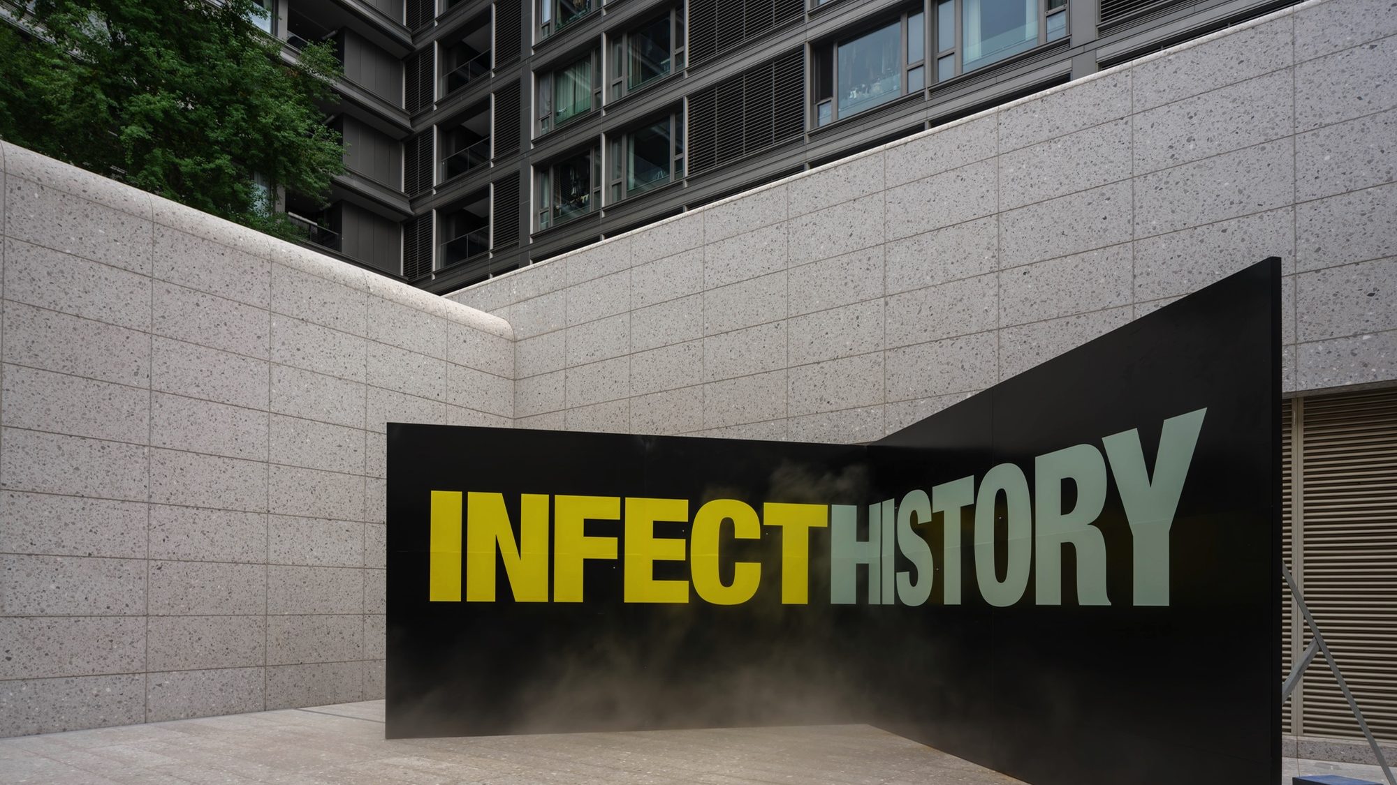 A black wall printed INFECT HISTORY, with mist drifting across the letters, in an outdoor courtyard