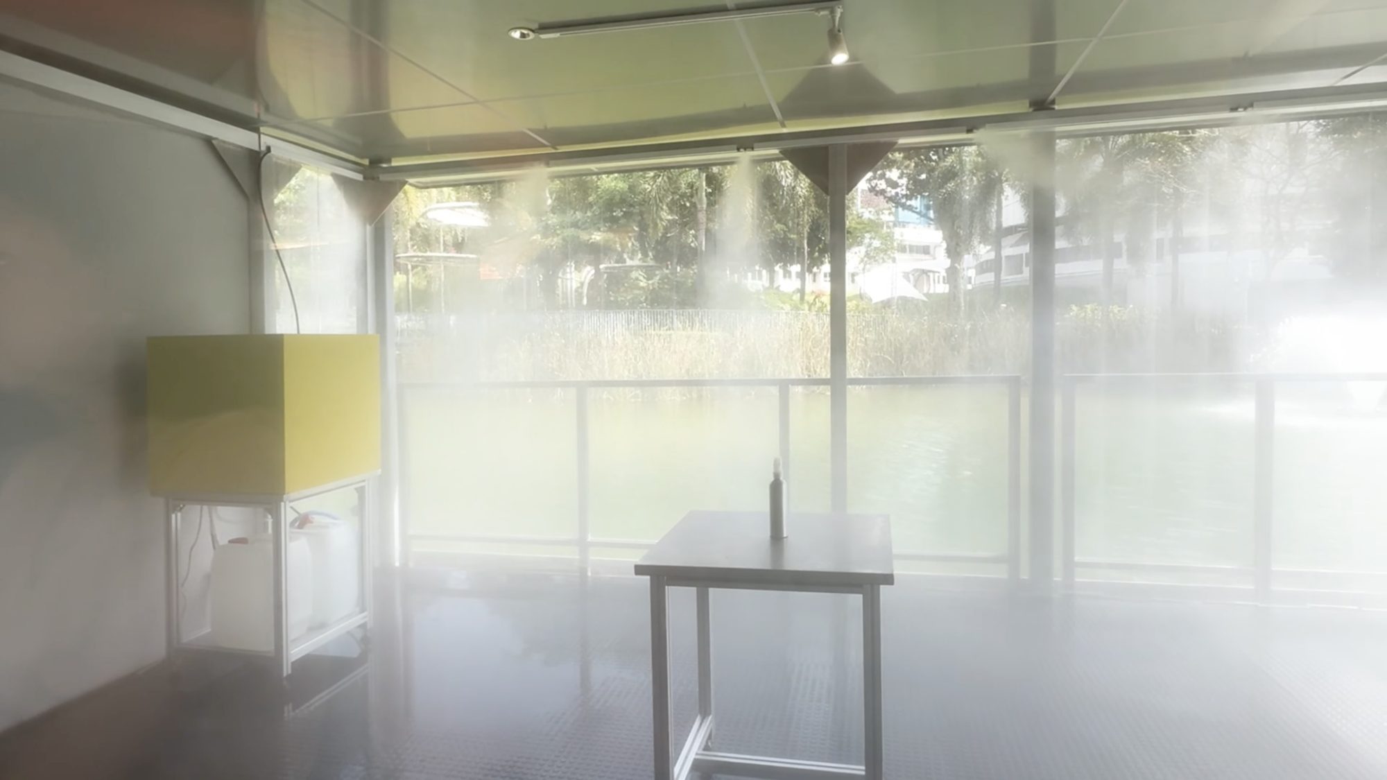 Inside the glass room, filled with mist, a yellow box and a table holding a bottle