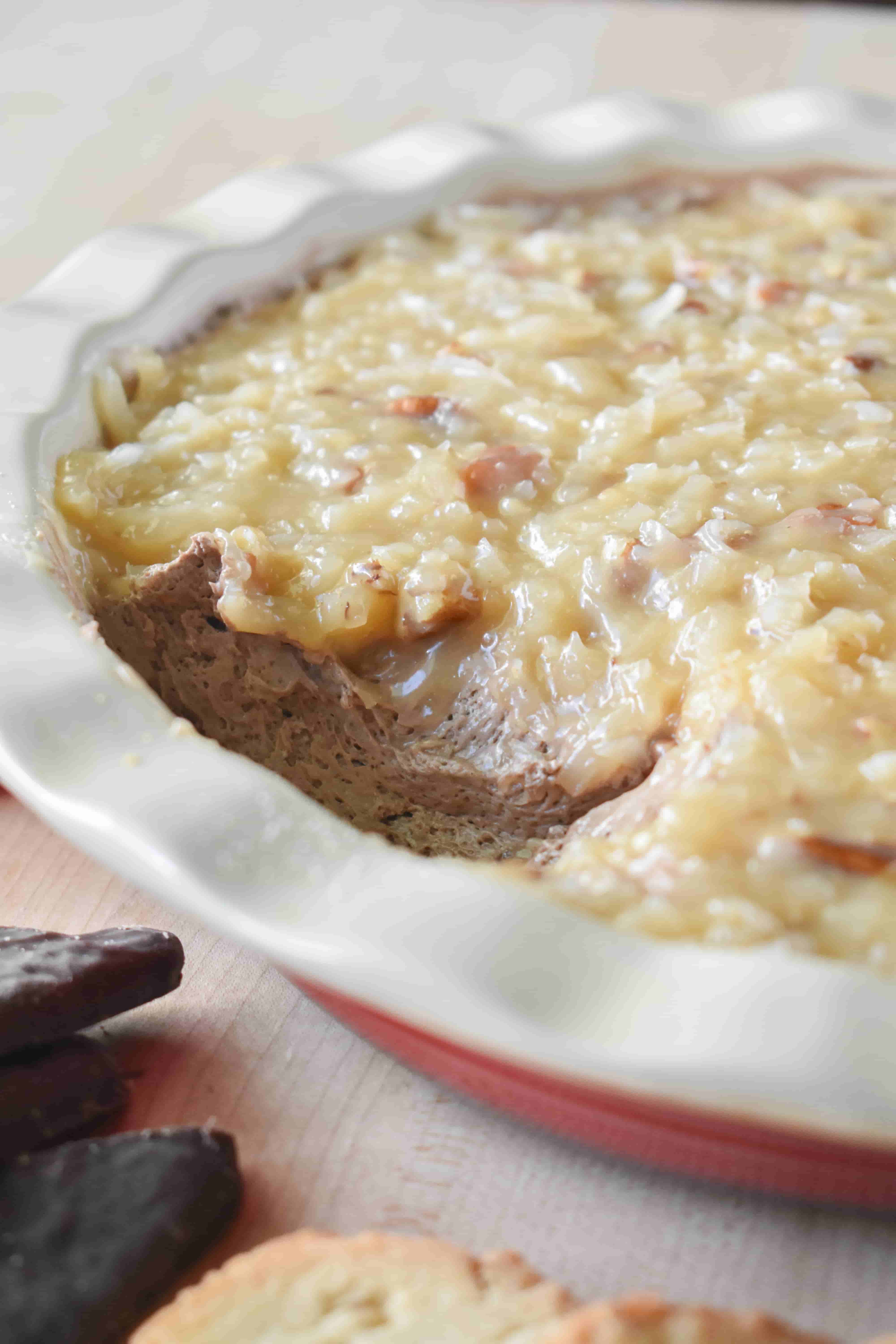 German Chocolate Dip in a red pie plate with a portion scooped out, showing the creamy chocolate mascarpone base layered under the coconut pecan frosting topping, with dippers arranged around the dish.