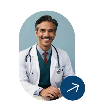 Smiling male doctor with stethoscope wearing a white coat and tie sitting with hands resting on a table against a blue background.