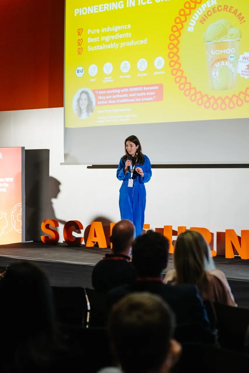 Woman with curly dark brown hair wearing glasses, a black and white patterned top, and a white blazer, smiling slightly.