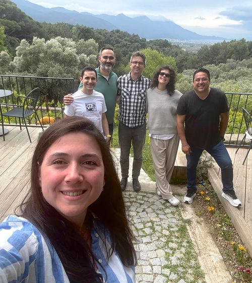 Group selfie of six smiling adults on a stone-paved patio with green trees and distant mountains in the background.