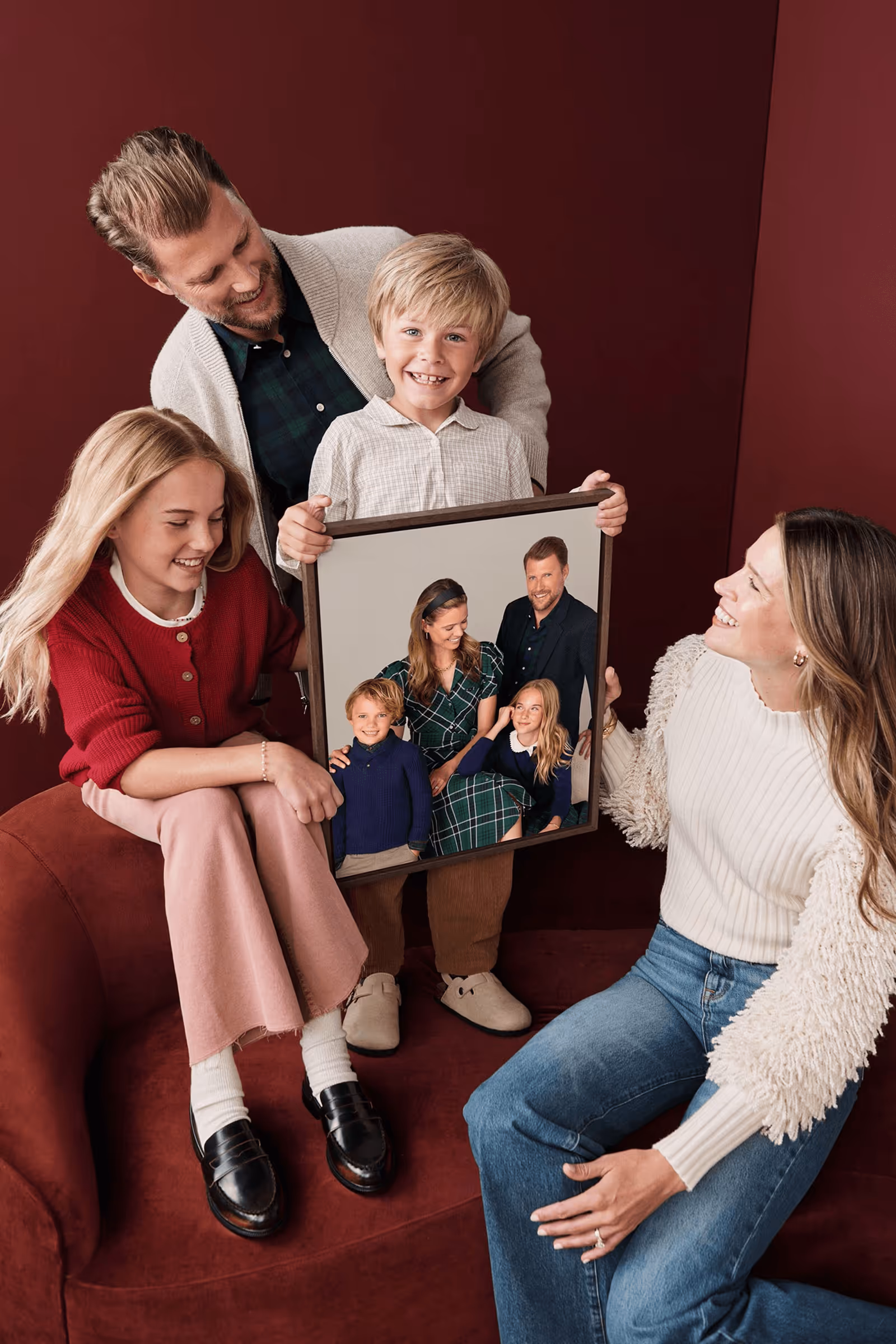 A smiling family of four sitting and standing on a red couch, with the boy holding a framed family portrait of the same group dressed formally.