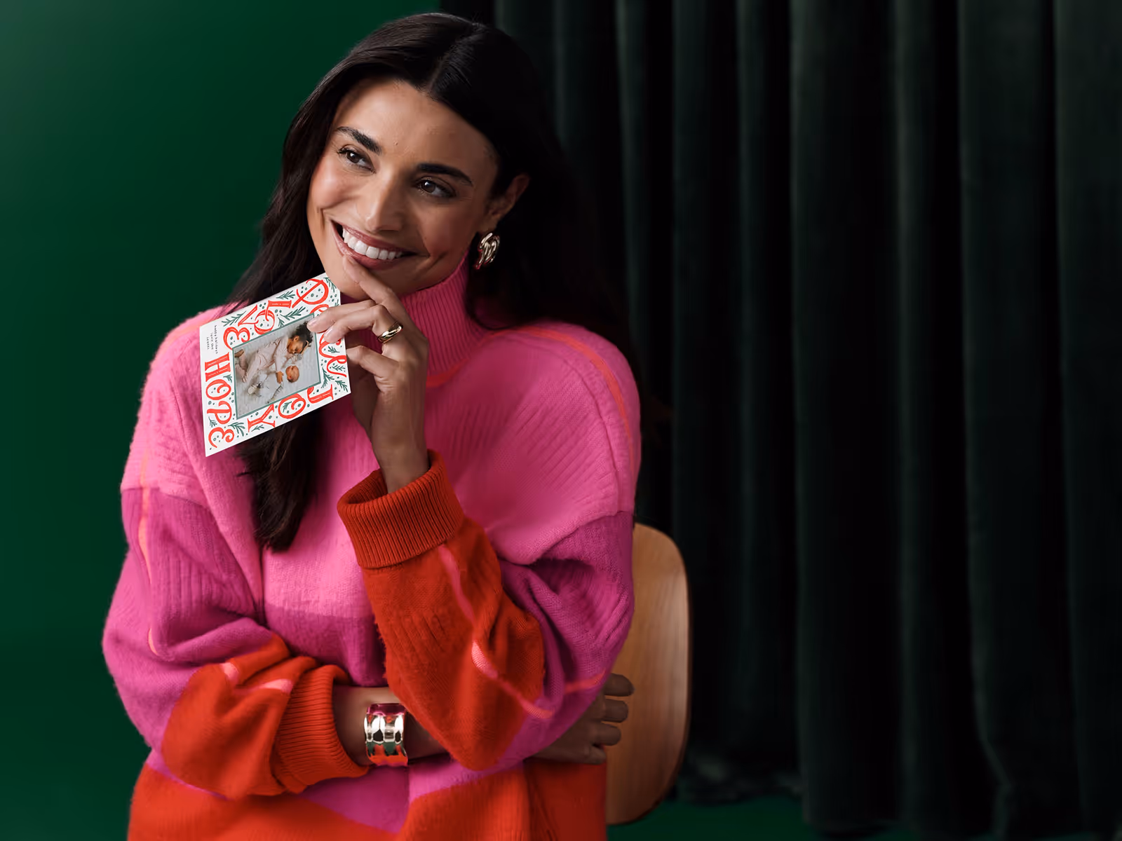 Smiling woman in a pink and red sweater sits holding a holiday card with a photo of two babies.