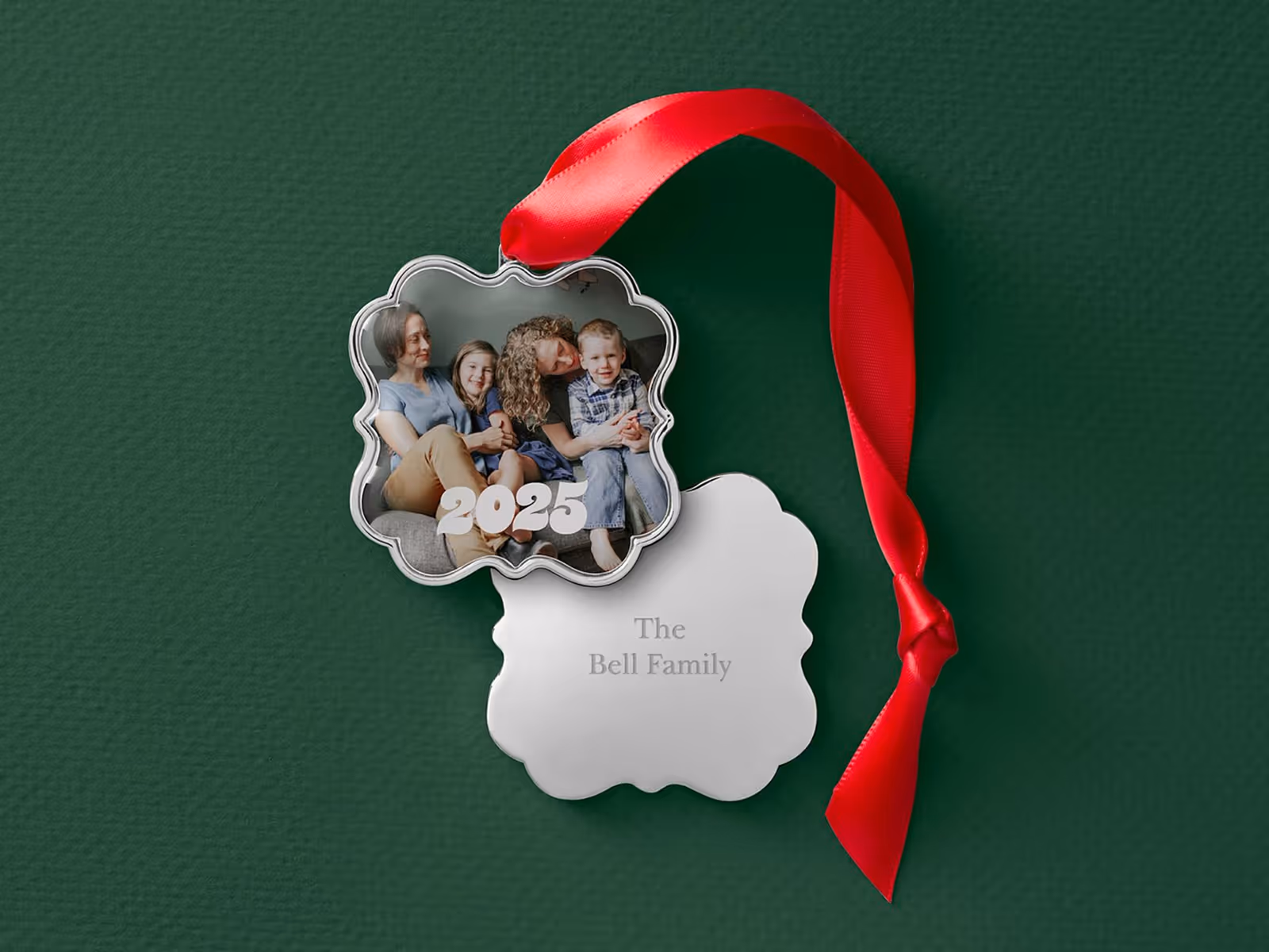 Scalloped silver holiday ornament with a family photo and the year 2025, attached to a red ribbon on a green background.