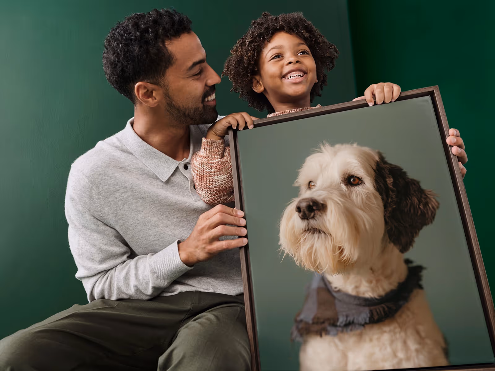 Smiling man and child holding a large framed portrait of a white and brown dog wearing a scarf.