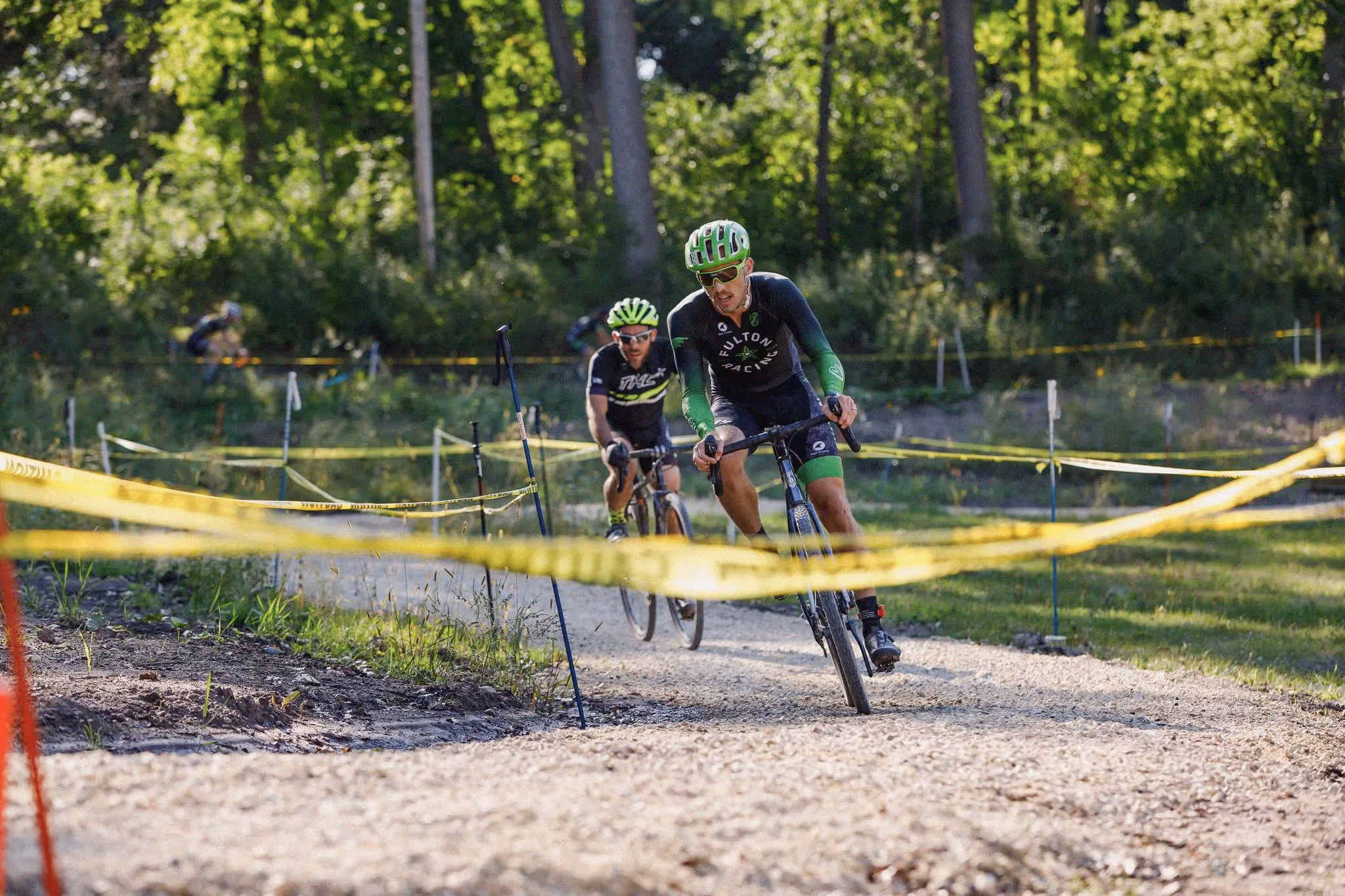 Two men wearing helmets racing bicycles on a gravel trail marked by yellow tape with a green forest background.