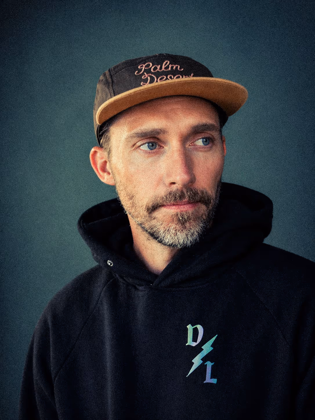 Bearded man wearing a black hoodie with colored letters and a lightning bolt, and a black and brown cap reading 'Palm Desert.'