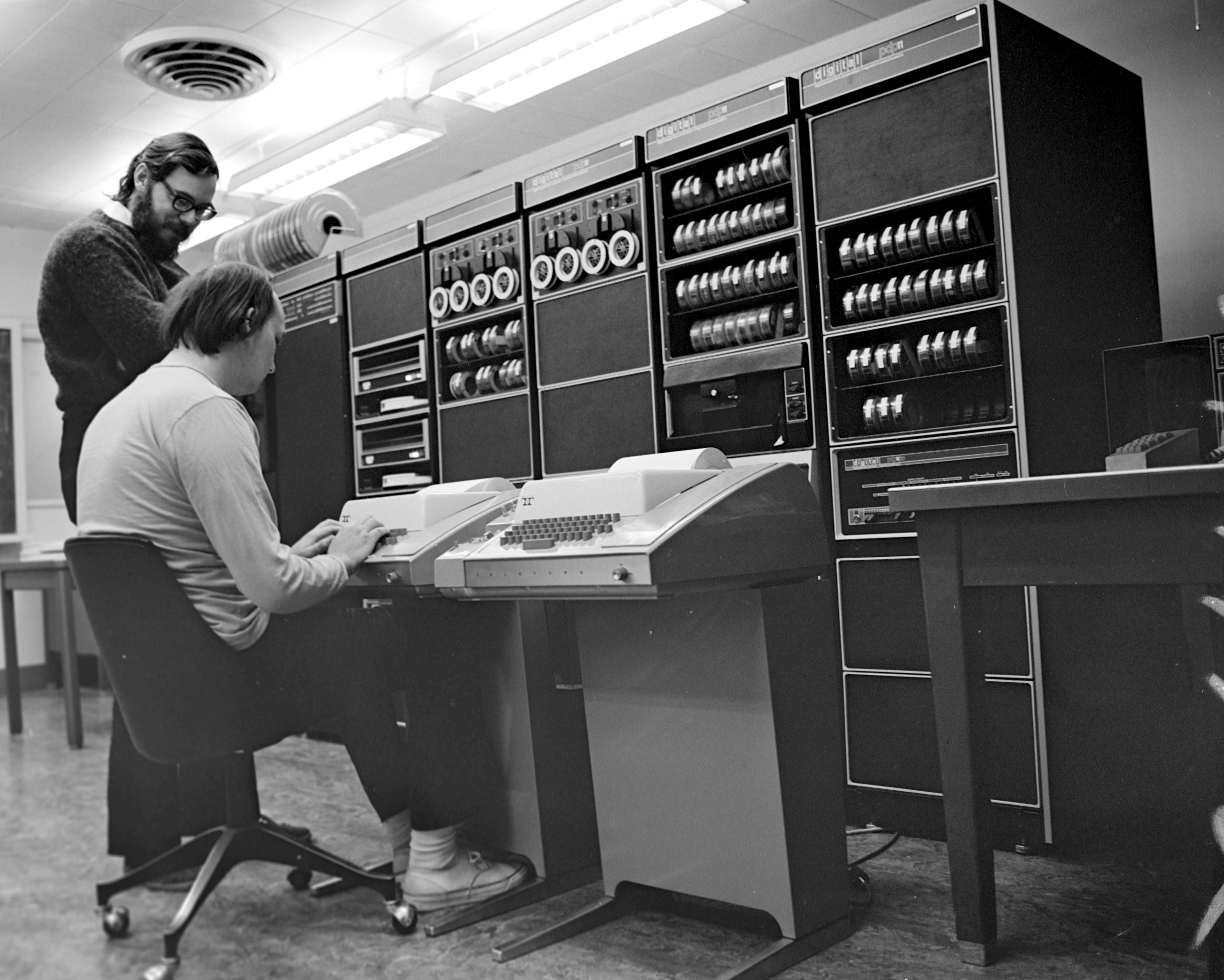 Dennis Ritchie (standing) with Ken Thompson (steated) in front of a large early computer.