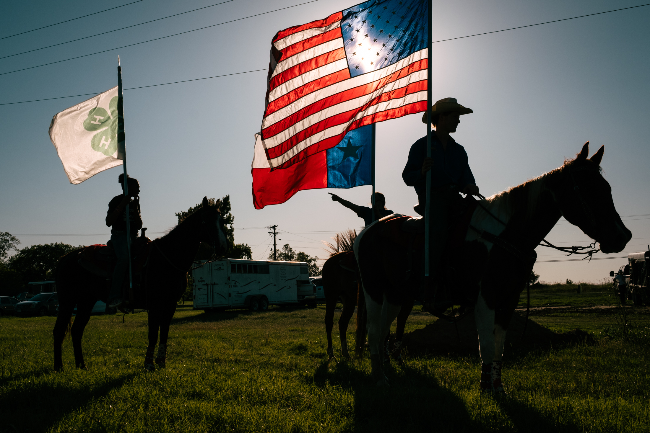As the sun sets behind them, three children on horseback practice carrying flags into the rodeo arena before the event starts. The children are carrying American, Texan, and 4H flags.