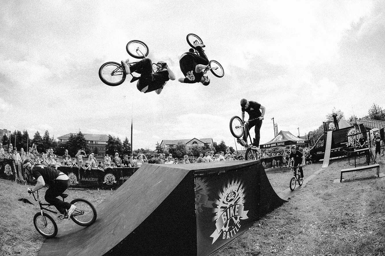 Sequence of a BMX rider performing a backflip over a ramp during an outdoor bike battle event with spectators in the background.
