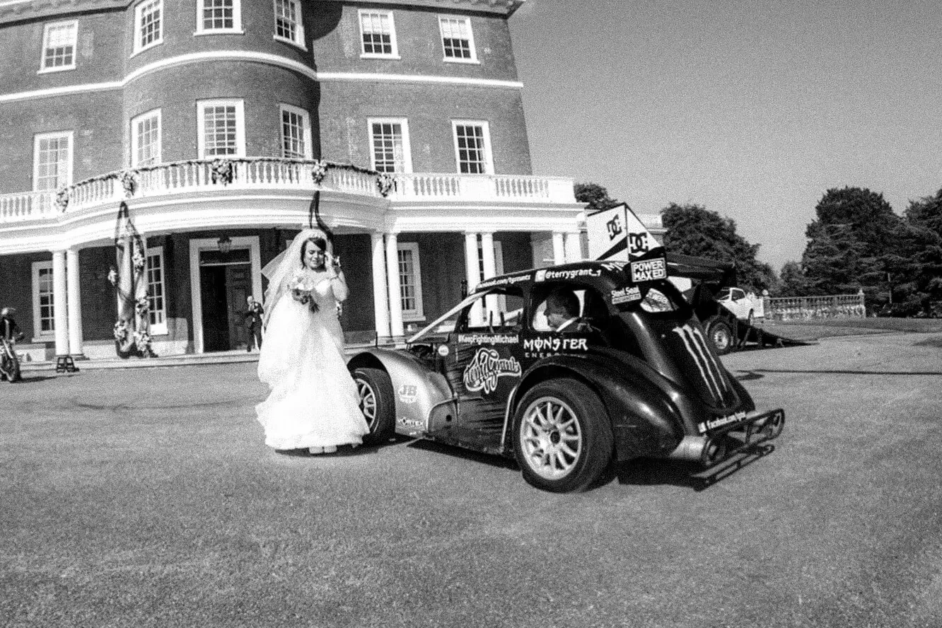 Bride in a wedding dress standing next to a black race car decorated with Monster Energy and DC logos outside a large brick mansion.