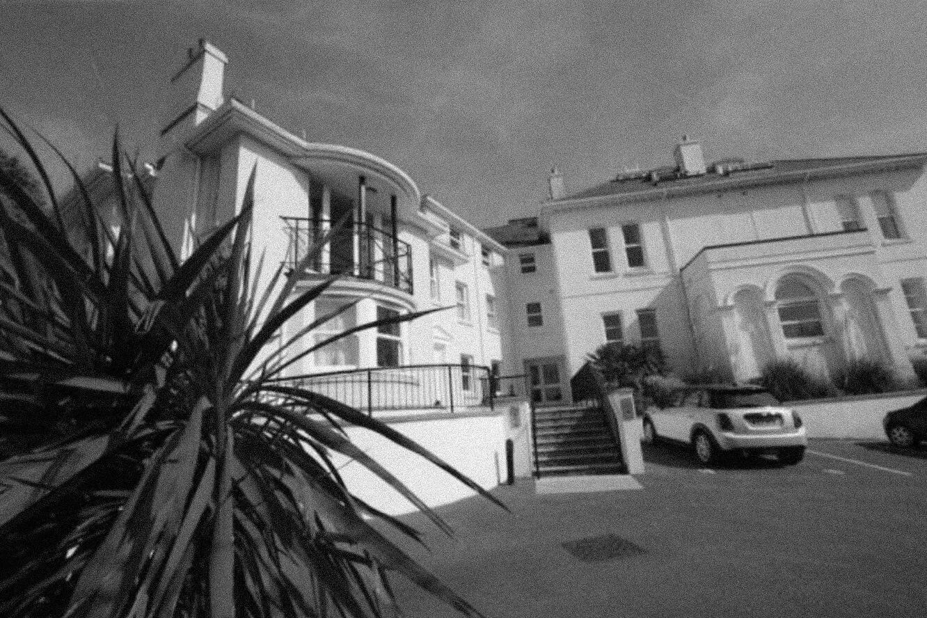 Exterior view of a large white building with curved balconies, stairs, and parked cars in front.