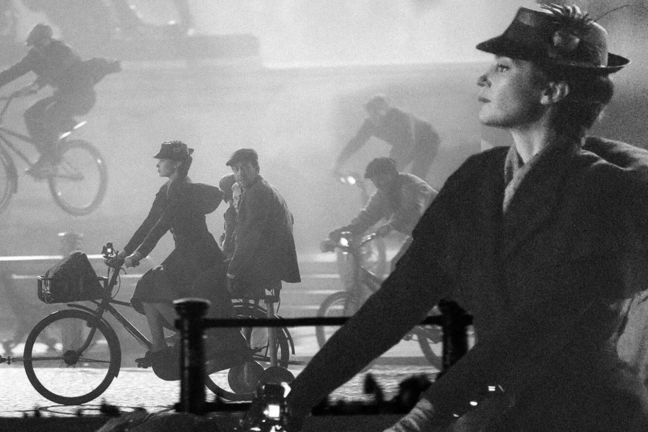 Black and white scene of people riding bicycles, including a woman in a hat in the foreground and others cycling in the background.