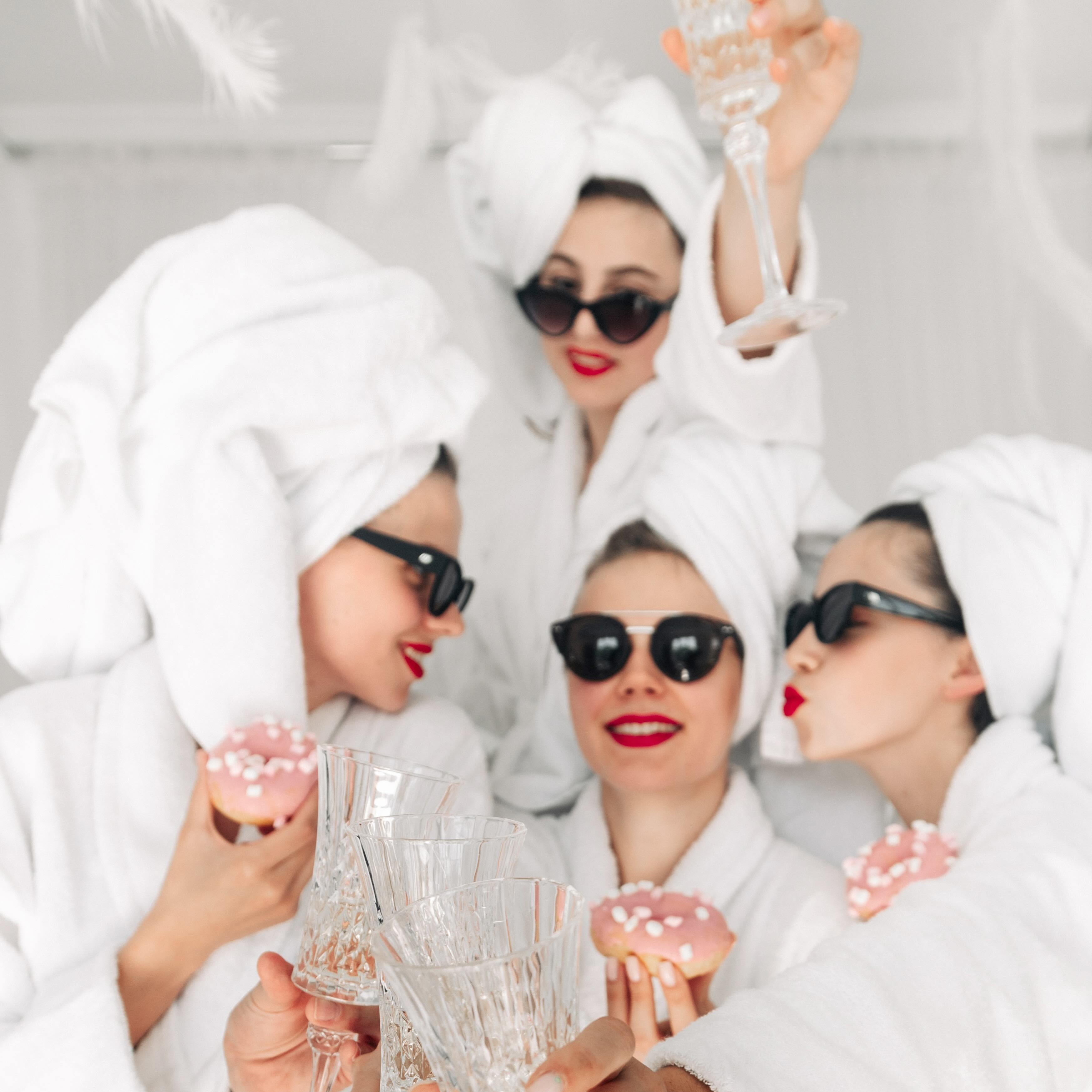 Four friends wearing white robes, towel wraps, and sunglasses raise their glasses together at a bachelorette party.