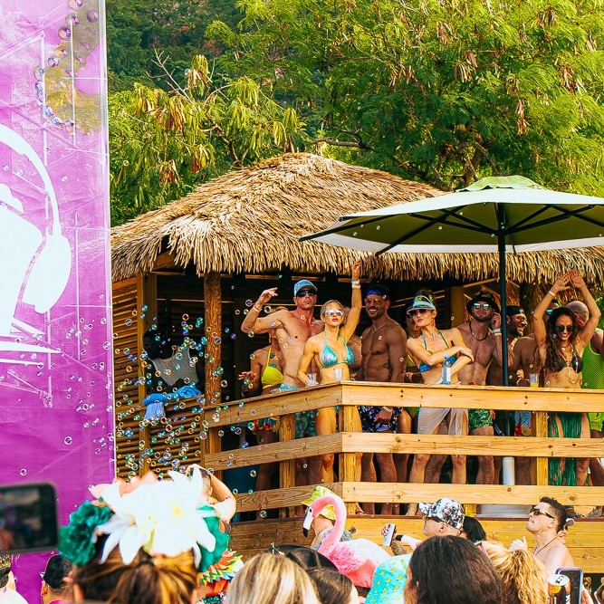 Friends celebrate on a shaded wooden cabana deck overlooking a Groove Cruise destination party.