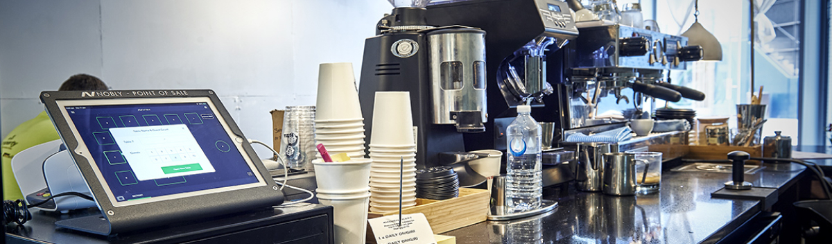 Counter of a modern coffee shop with a point-of-sale touchscreen device, stacks of disposable cups, a water bottle, and a professional espresso machine in the background.