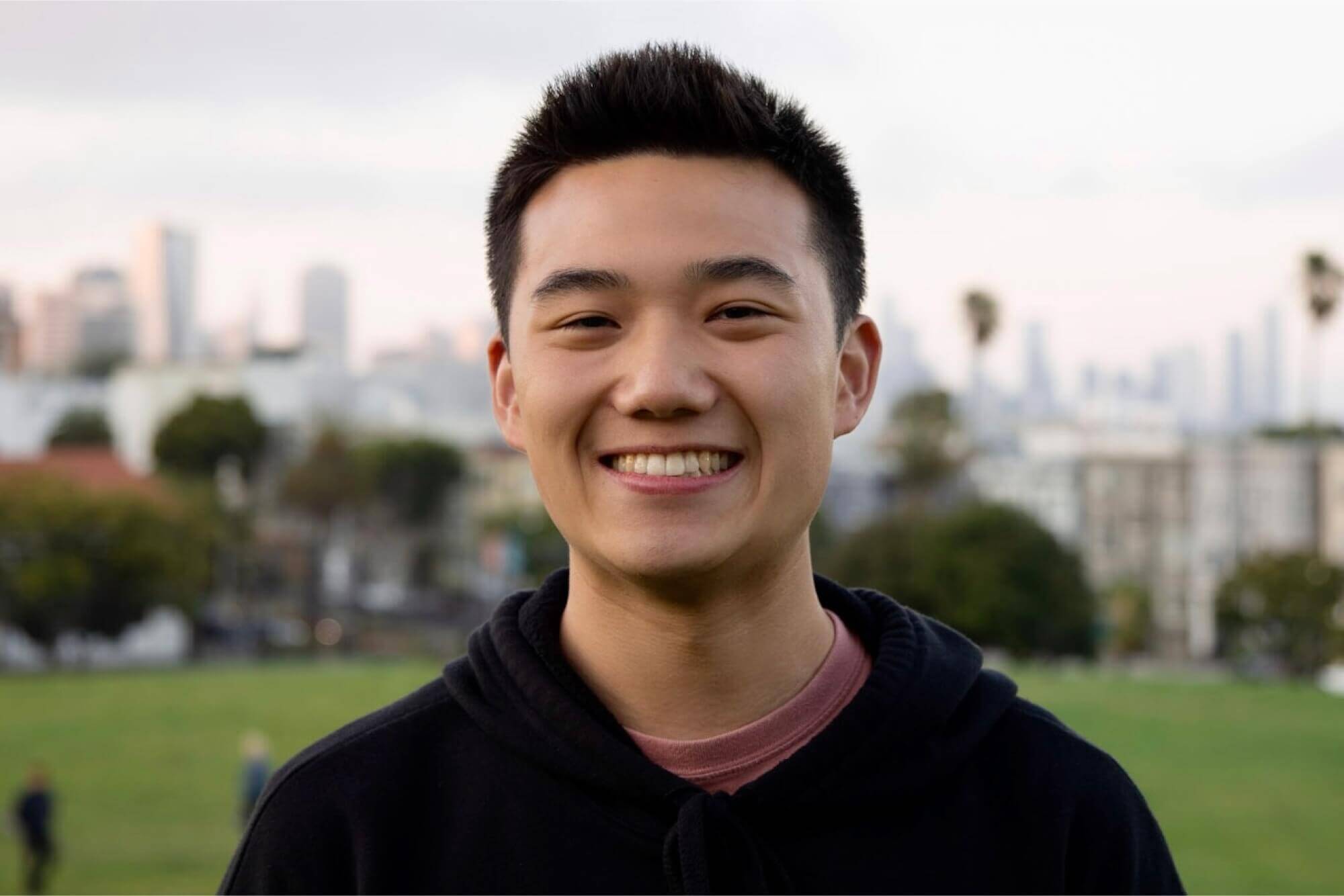 Austin Lee, smiling in a black hoodie, stands in a park with a blurred city skyline behind him.