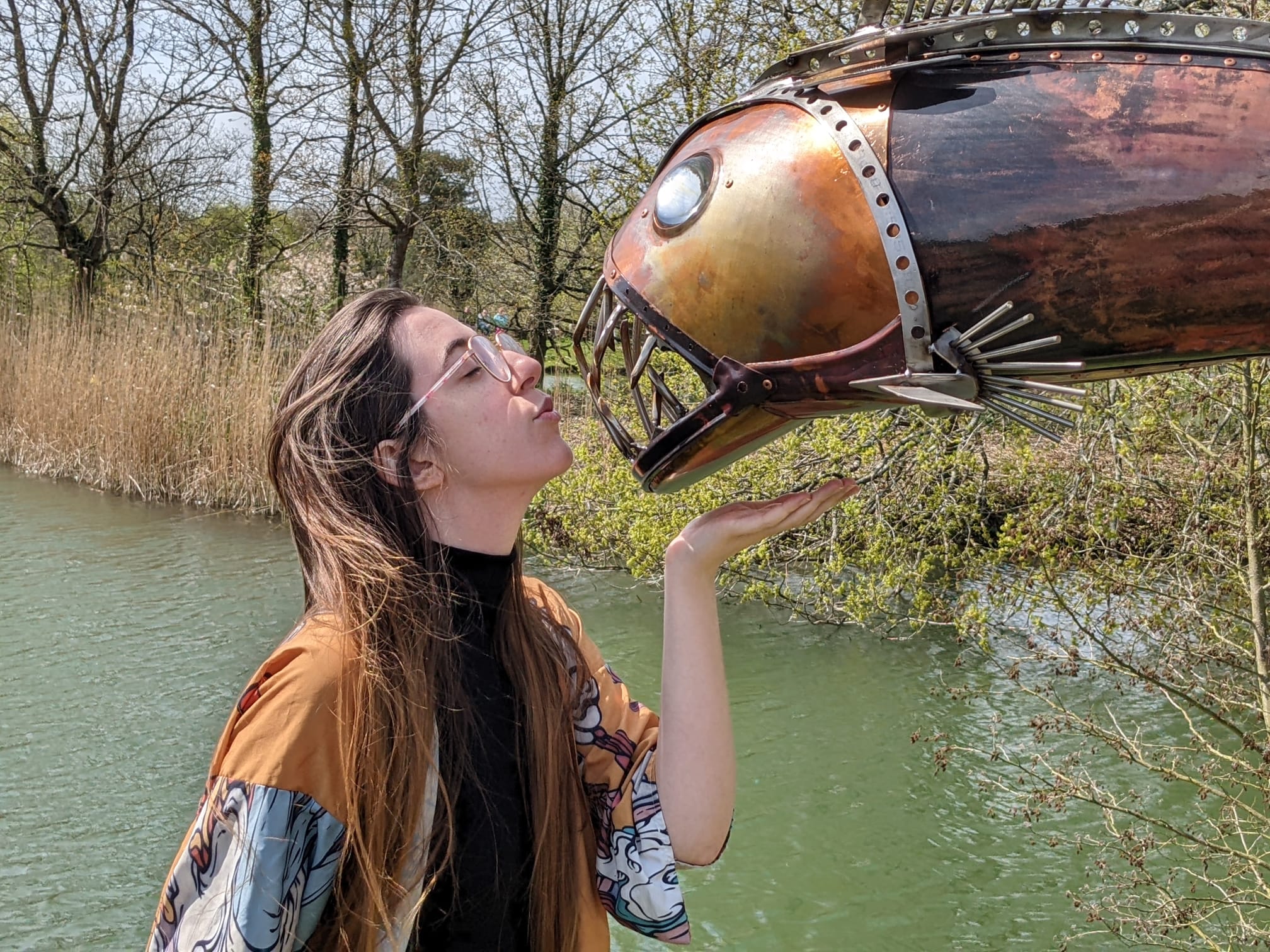 An image of me, a white woman with brunette, long hair. She's in front of a statue of a fish that's made out of recycled metal.