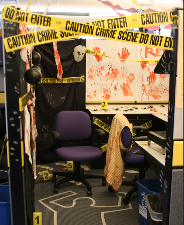 A cubicle decorated as a crime scene with a white body outline on the carpet and crime scene tape.