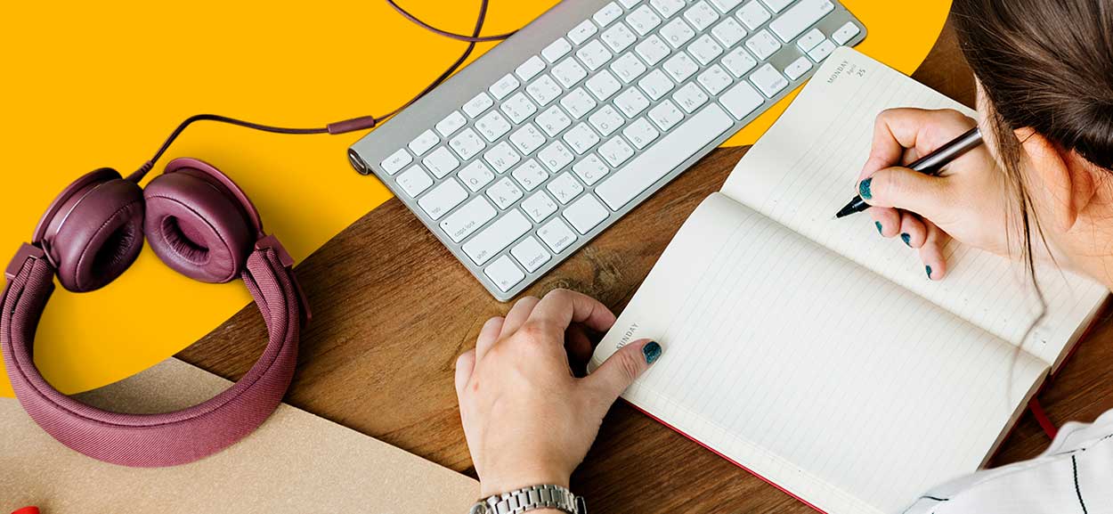 A designer writes notes in her notebook. A keyboard and a set of headphones sit in her workspace.