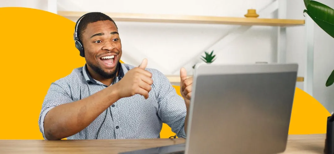 A man giving two thumbs up as he works on a laptop. He is sitting against a yellow Designity blob background.