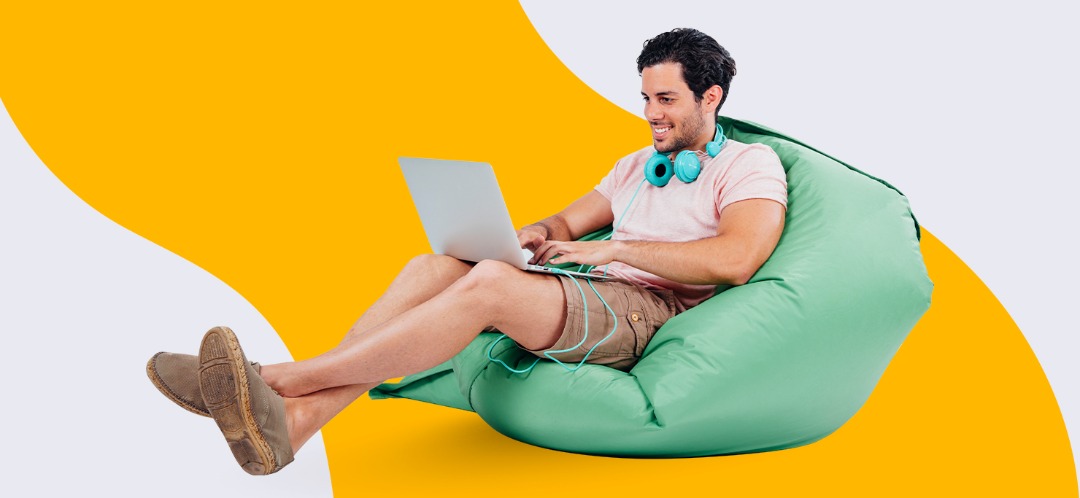 A man in shorts lounges on a beanbag and works on a laptop, against a white and yellow Designity background.