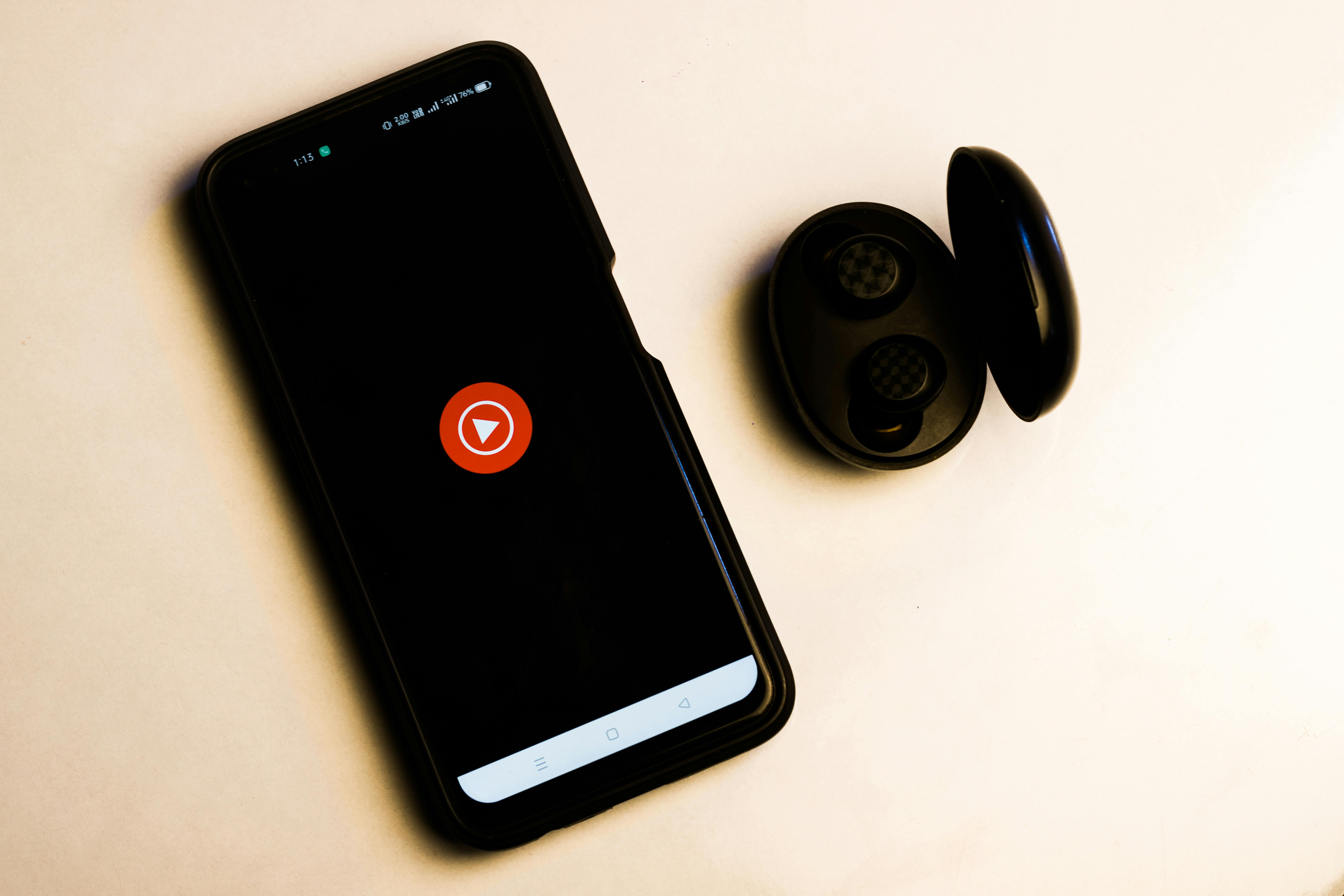 A smartphone displaying a red play button on its screen lies next to an open black wireless earbuds case, both placed on a light-colored surface.