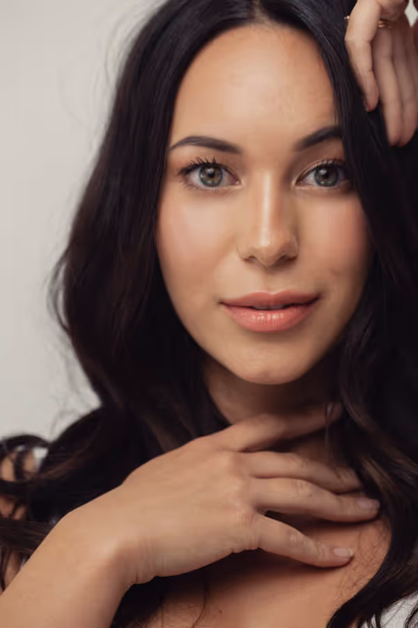 Headshot of a dark-haired woman with clear skin and one hand on her collarbone