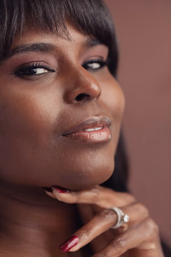 Close-up of a woman’s face with bangs, clear brown skin, and her hand under her chin