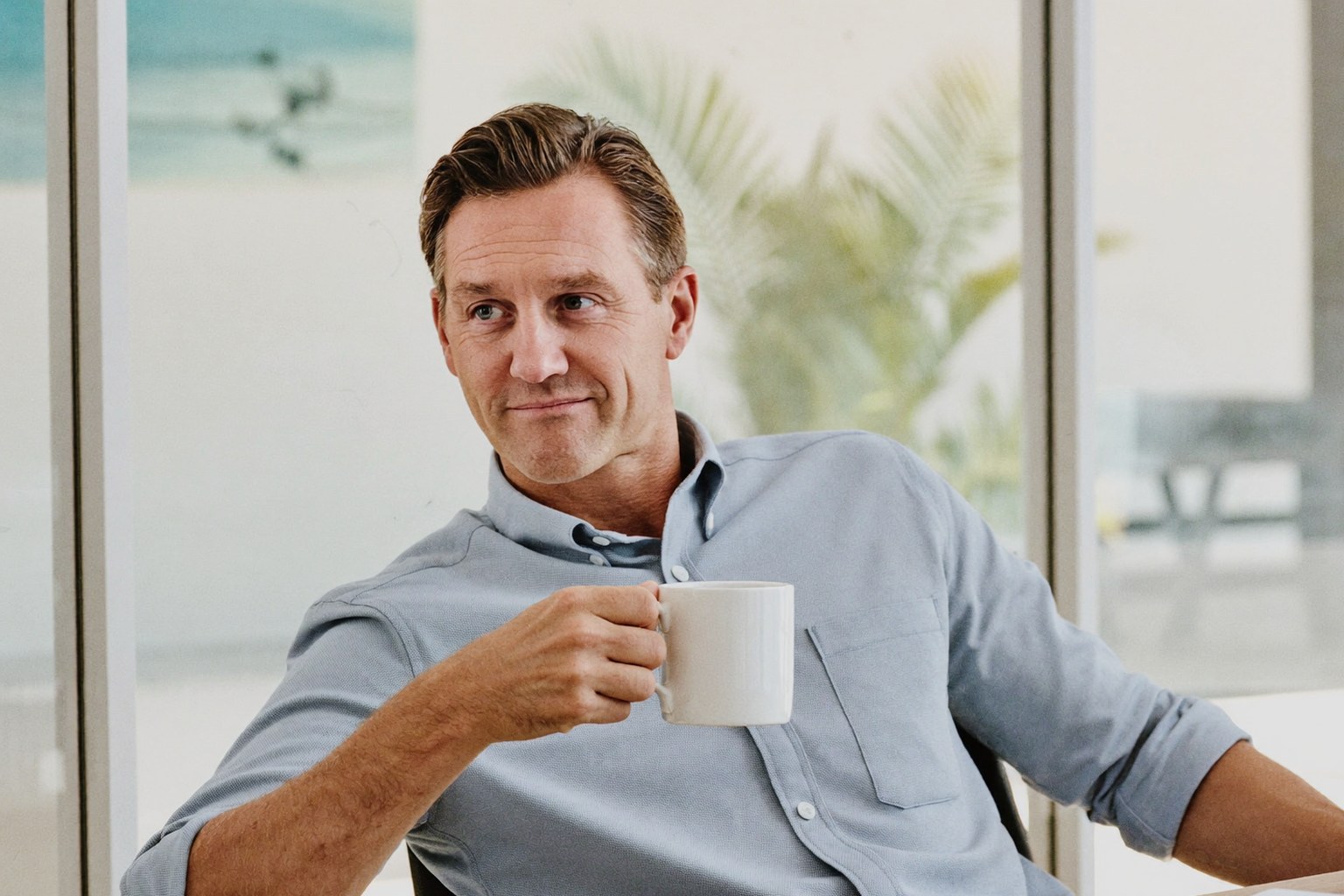 Finance leader in a light blue shirt relaxing with a coffee in a bright glass-walled meeting room