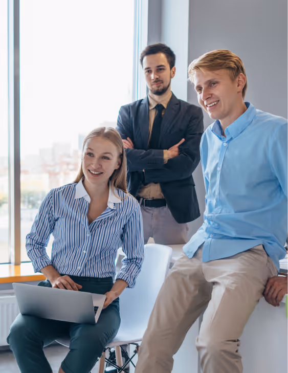 Three young professionals, two seated and one standing, smiling in an office with large window.