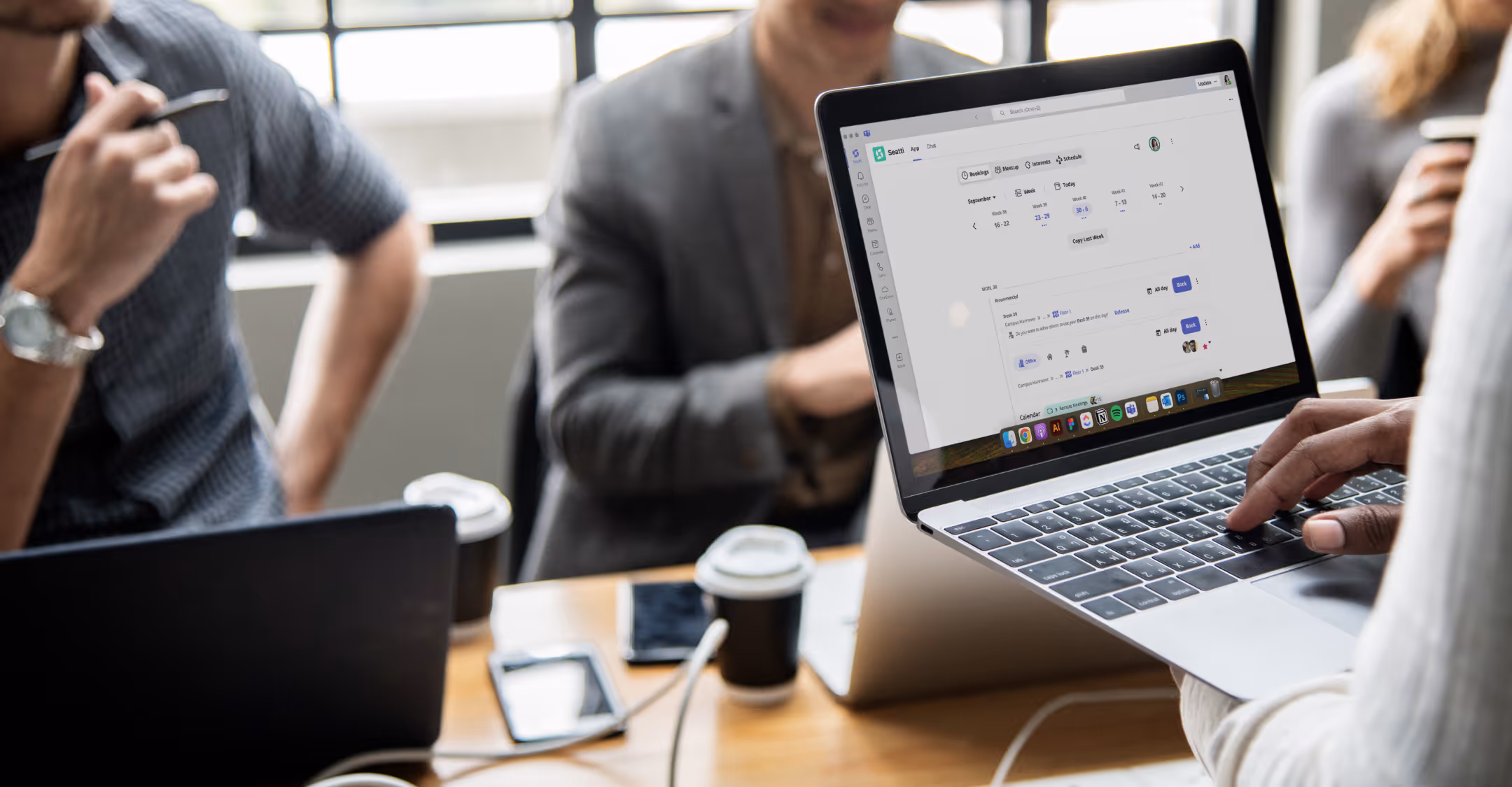 Person holds laptop showing a desk booking app, others sit at table with laptops and coffee cups.