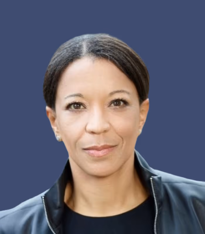 Porträt einer Frau mit zurückgekämmtem dunklem Haar und schwarzem Jackett vor blauem Hintergrund.