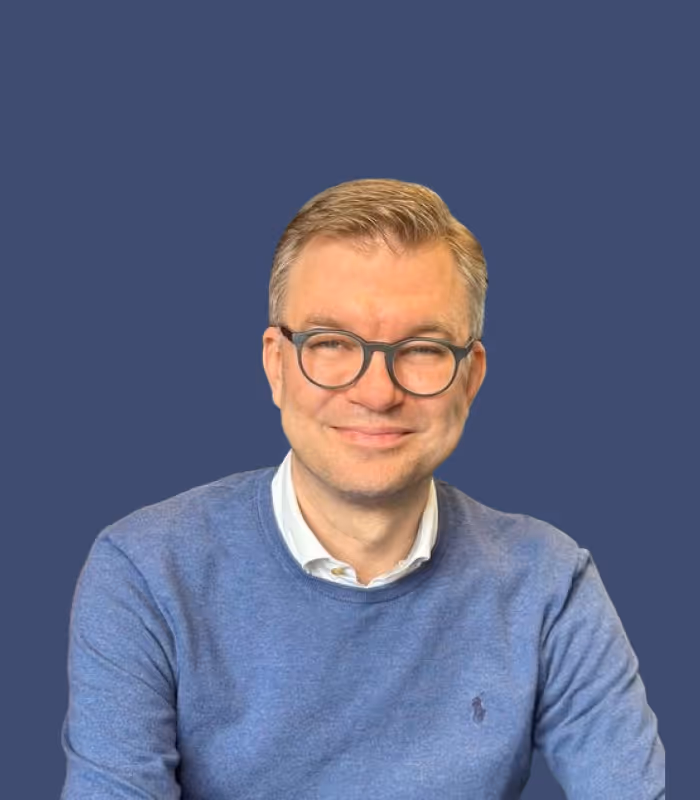 Mann mit Brille und blauem Pullover lächelt vor dunkelblauem Hintergrund.