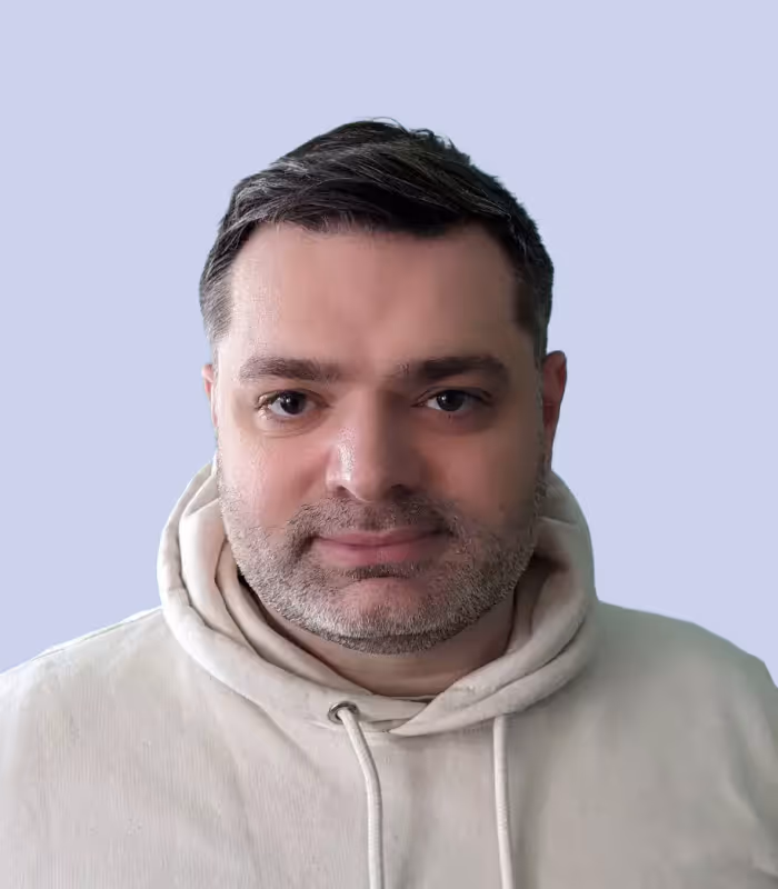 Porträt eines Mannes mit kurzem Haar und grau meliertem Bart in einem hellen Kapuzenpullover vor blauem Hintergrund.