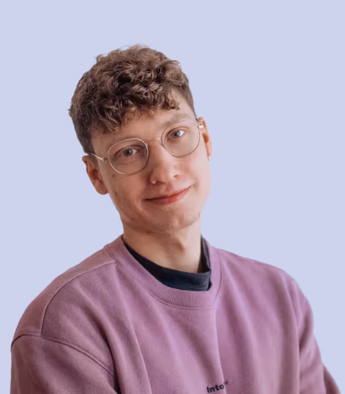 Junger Mann mit lockigem Haar, Brille und pinkem Pullover vor hellblauem Hintergrund.