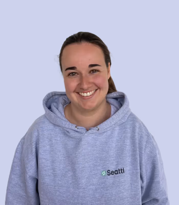 Lächelnde Frau mit braunen Haaren in grauem Kapuzenpullover mit Seatti-Logo vor hellblauem Hintergrund.