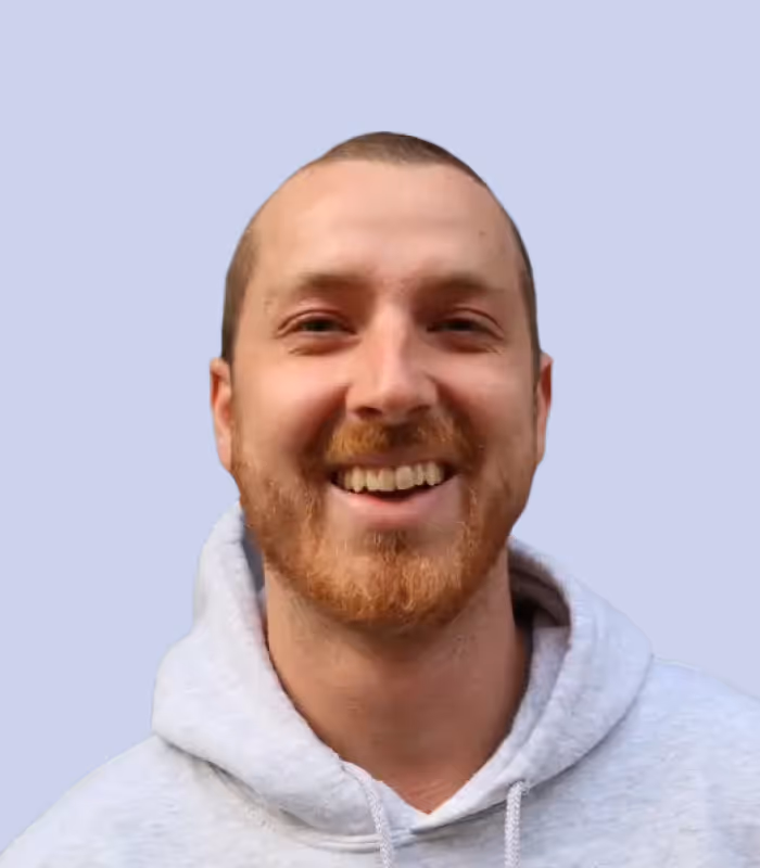 Junger Mann mit rotem Bart und Kapuzenpullover lächelt vor blauem Hintergrund.