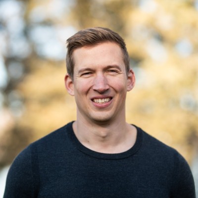 Smiling young man with short light brown hair wearing a dark sweater standing outdoors with blurred trees in the background.