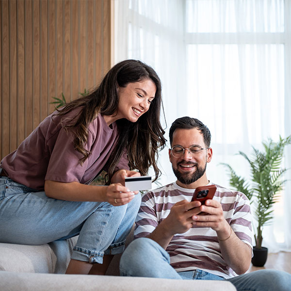 Hymyilevä pariskunta tekee yhdessä verkko-ostoksia älypuhelimella ja luottokortilla kotisohvalla.