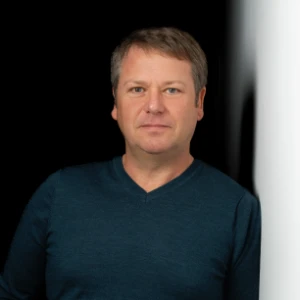 Man with short light brown hair wearing a dark blue shirt against a black and white background.